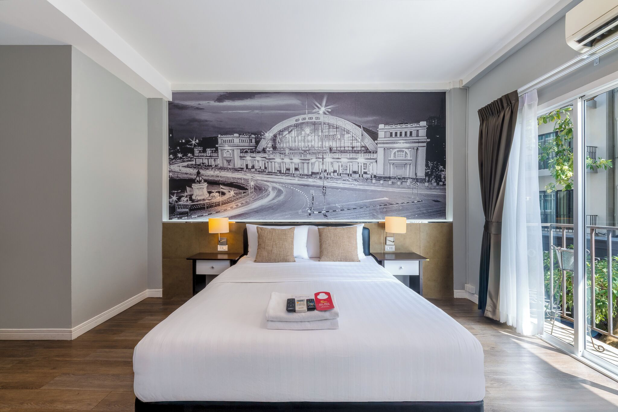 A hotel room with a large bed, two bedside tables with lamps, a large window with curtains, and a cityscape mural on the wall.