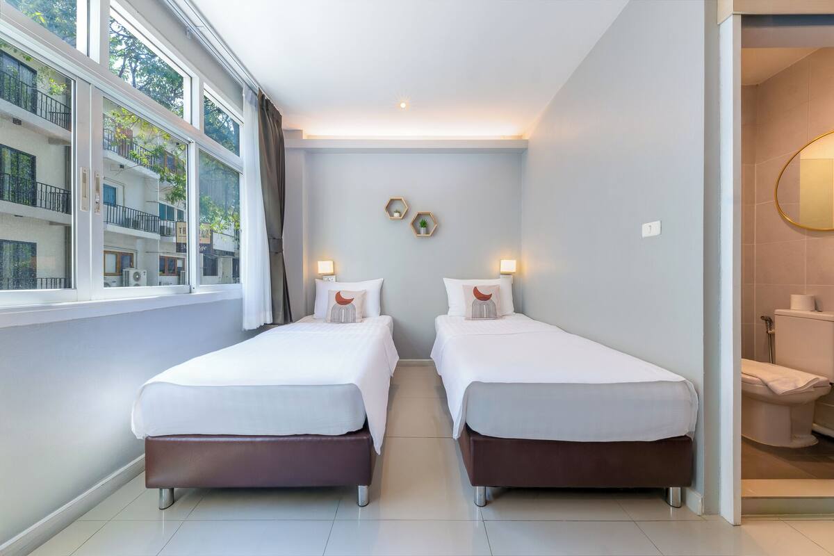 Quarto de hotel com duas camas, uma janela grande, vista de um edifício e banheiro visível por uma porta aberta.
