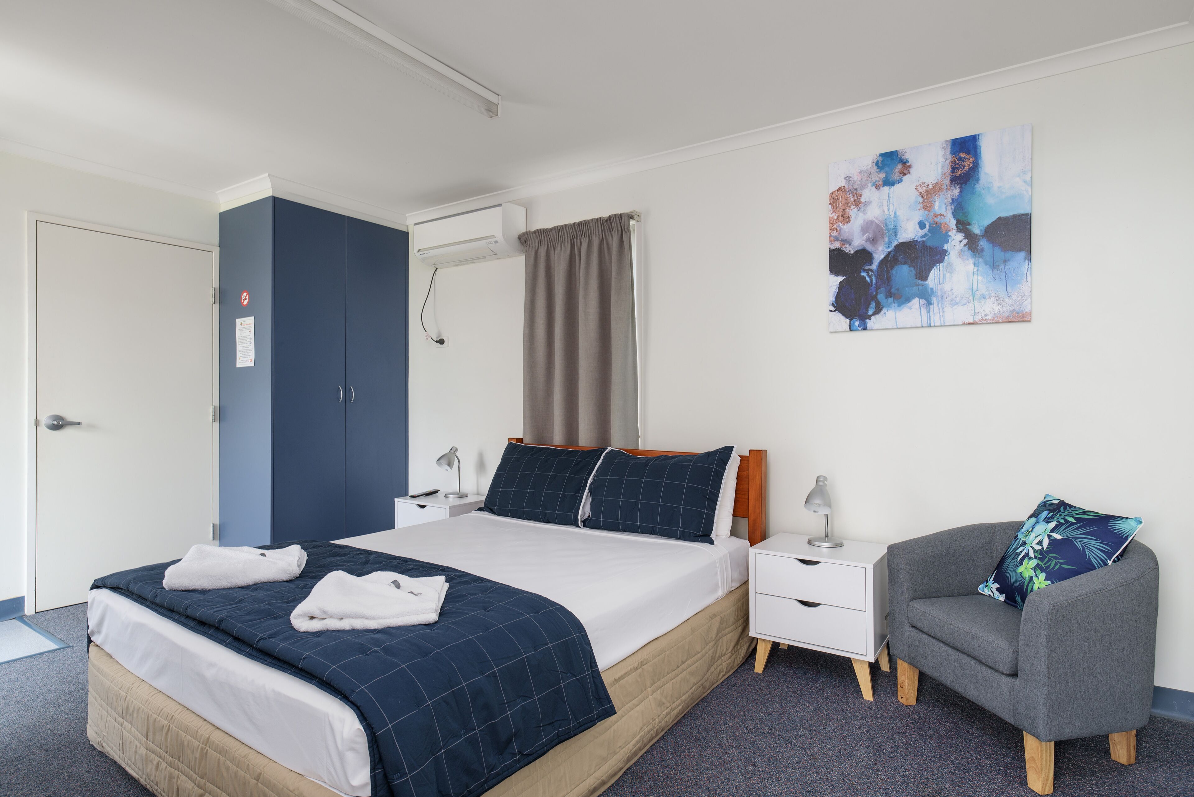 Ein Hotelzimmer mit Bett, Nachttisch, Stuhl, einem Bild an der Wand und einer Tür mit Klimaanlage.