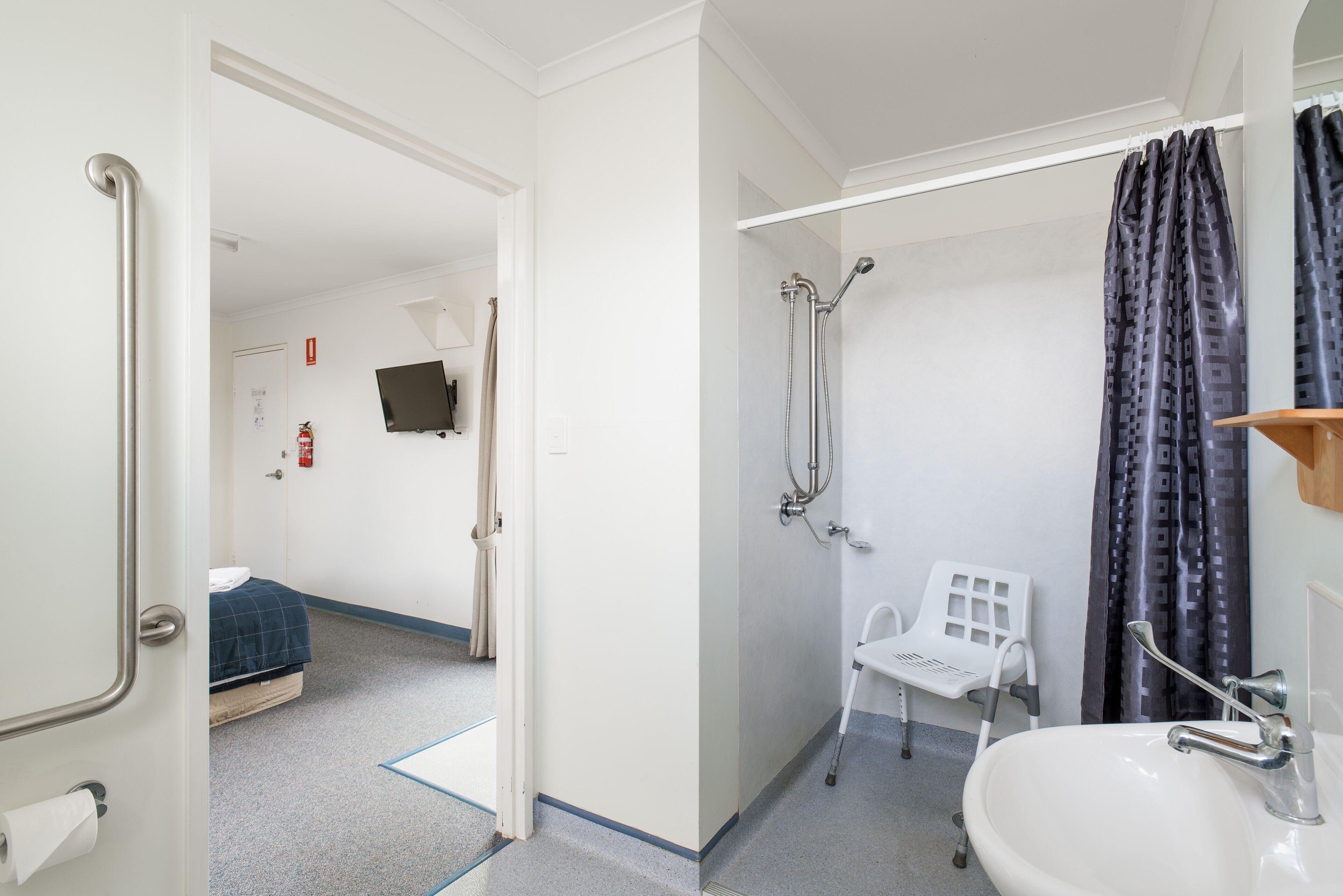 A hotel bathroom with a shower, sink, and a bed in the adjacent room.