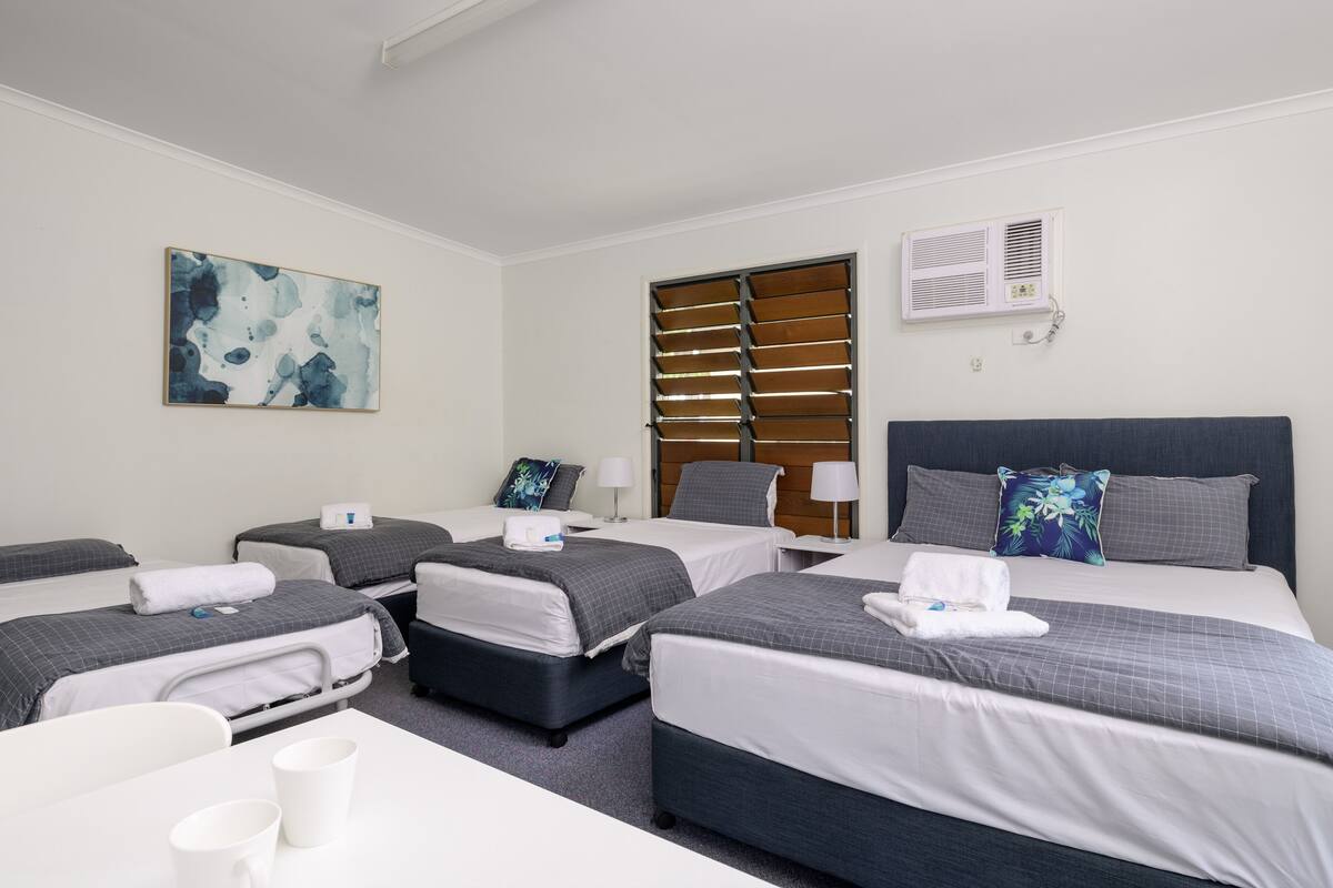 Une chambre d’hôtel avec quatre lits simples, un bureau et une chaise. Il y a un tableau au mur et un appareil de climatisation.