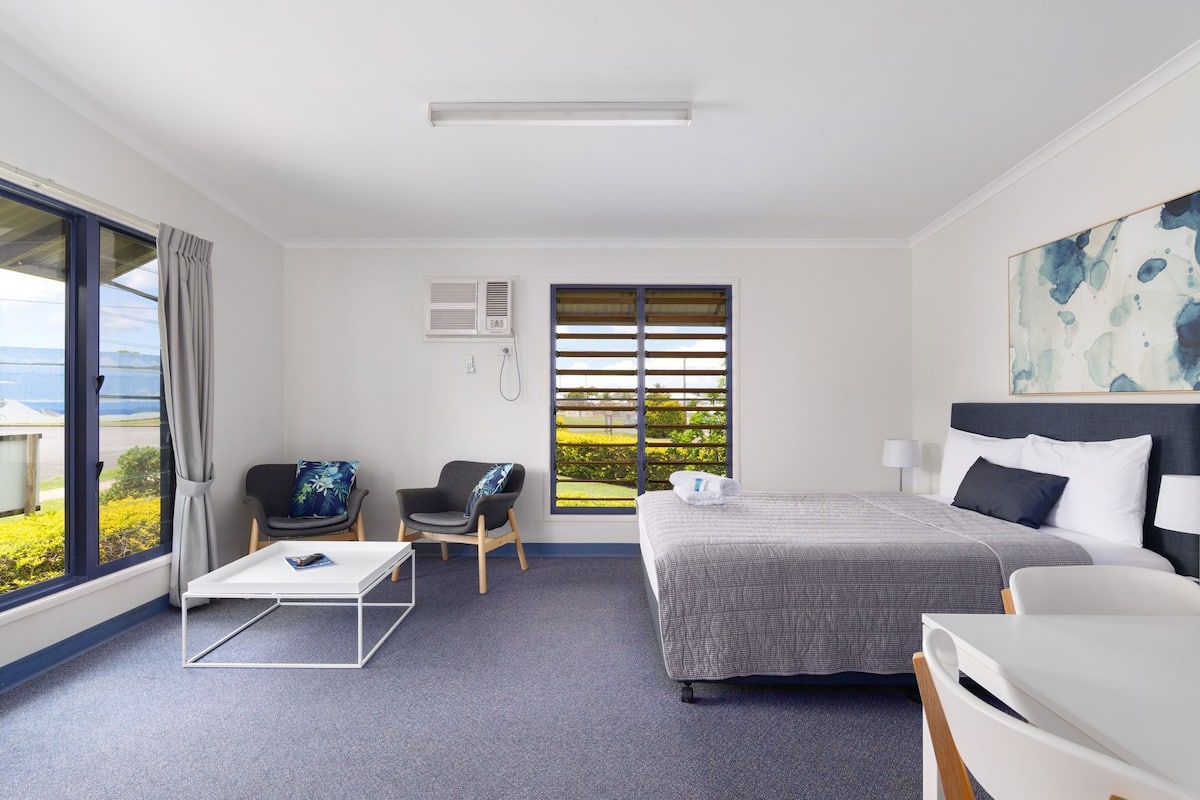 Une chambre d’hôtel comprenant un lit, un bureau, deux chaises, une fenêtre avec des stores, un appareil de climatisation et un tableau au mur.