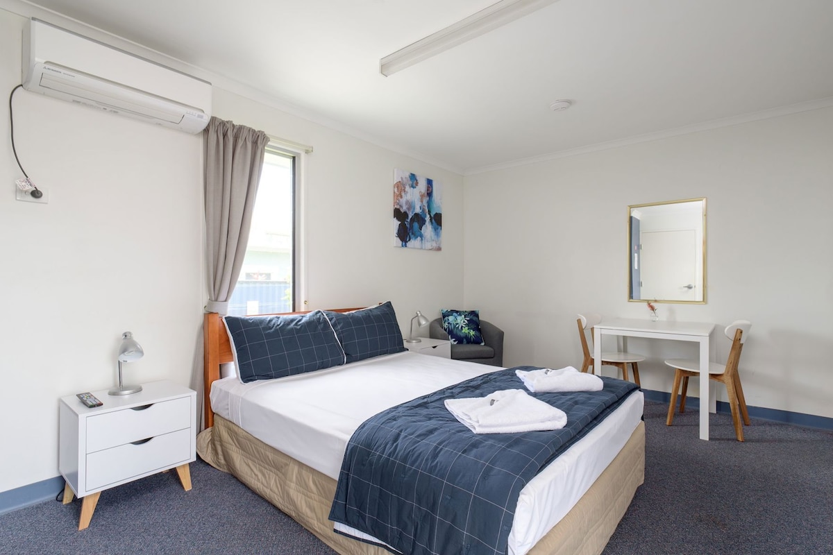 A hotel room with a bed, bedside table, desk, chair, and a window with curtains.