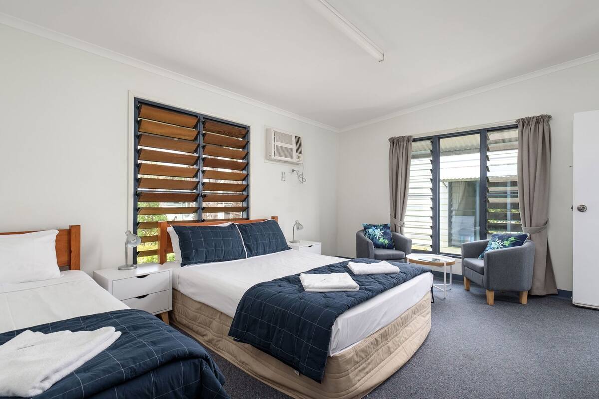 Une chambre d’hôtel avec deux lits, un bureau, une chaise et de grandes fenêtres équipées de stores.