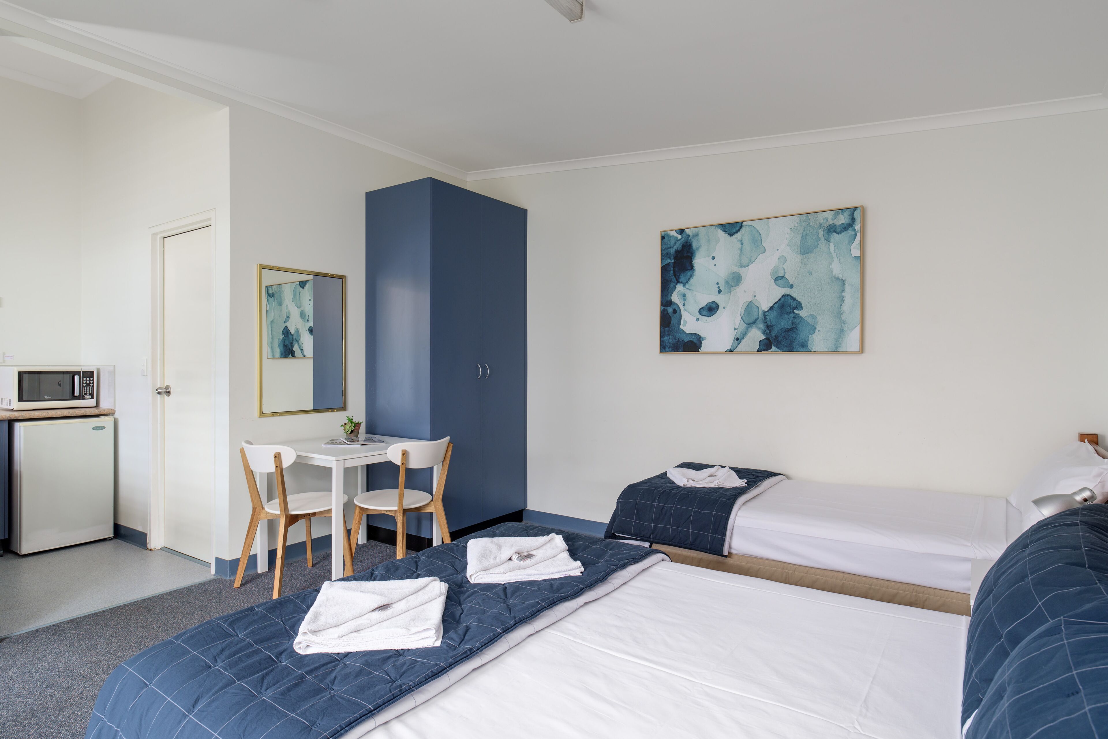 A hotel room with two beds, a desk, a chair, a microwave, and a painting on the wall.