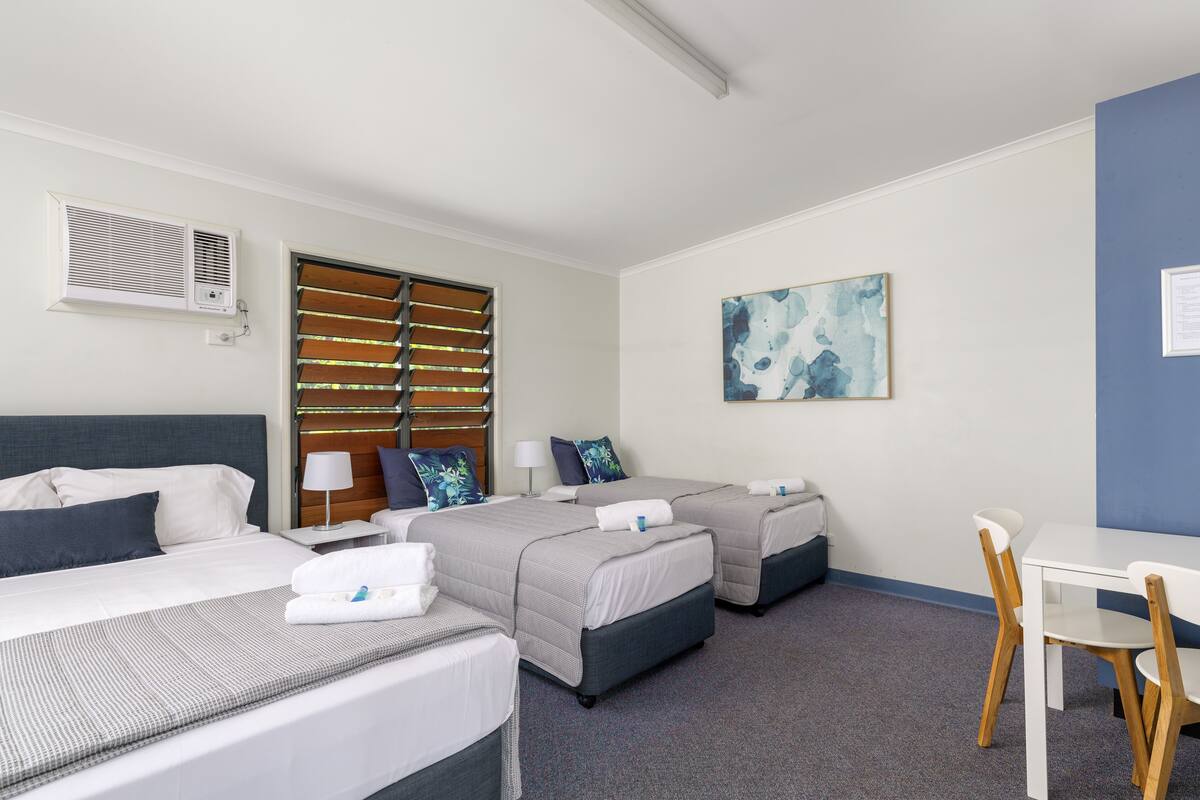 Une chambre d’hôtel avec deux lits, un bureau et une chaise. Il y a une fenêtre avec des stores, un climatiseur fixé au mur et un tableau accroché au mur.