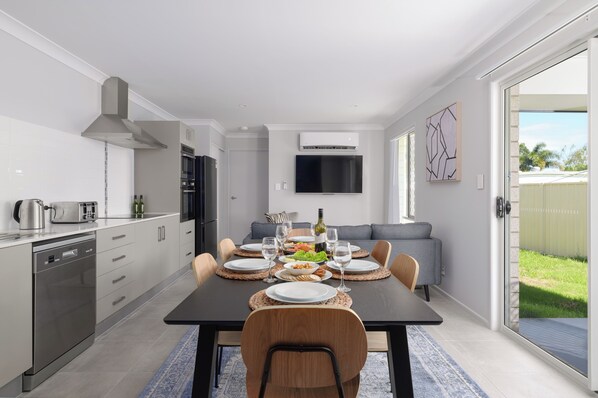 A modern kitchen with built-in appliances, a dining area with a table set for four, and a living area with a sofa and TV.
