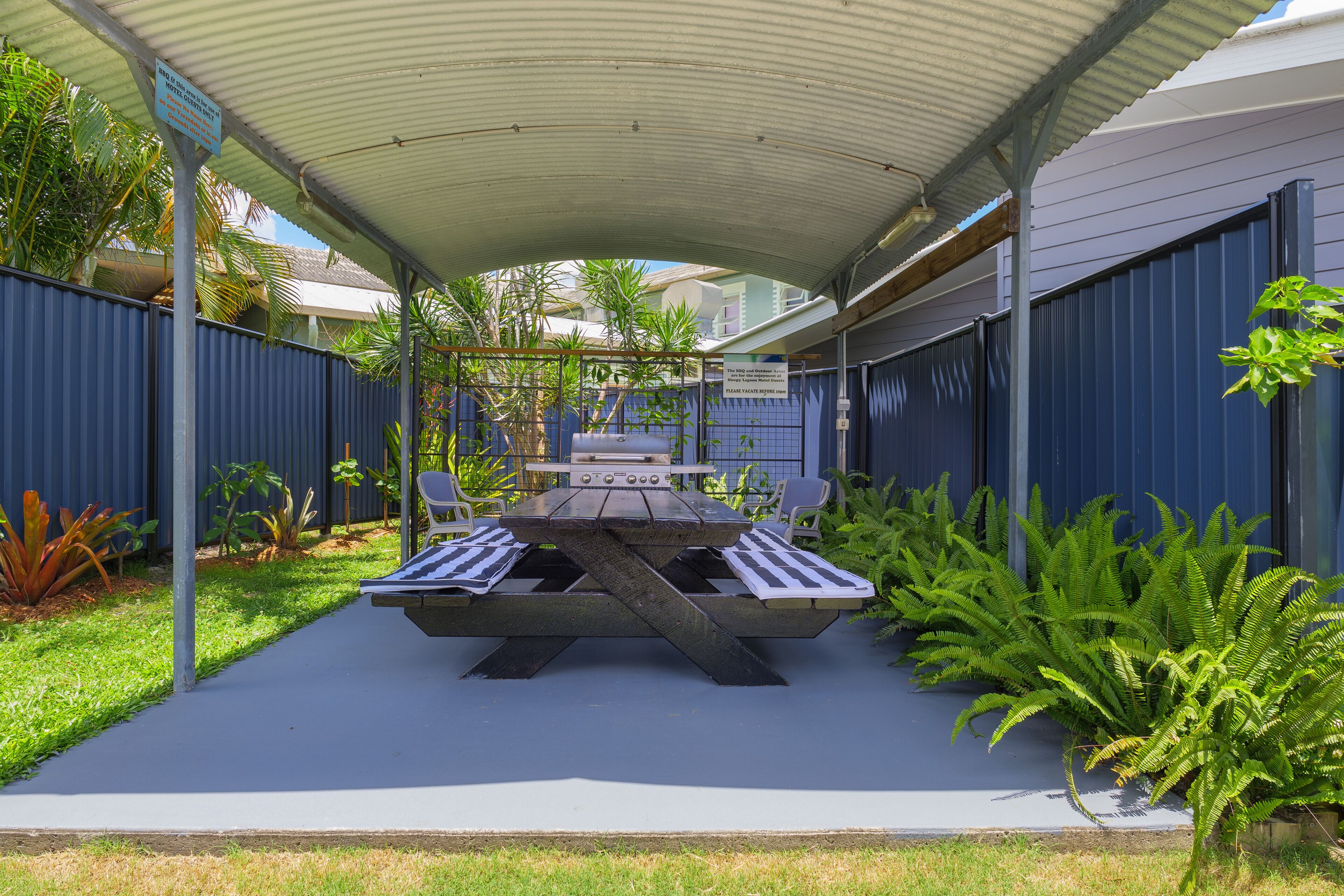 An outdoor patio area with a covered table and chairs, surrounded by a blue fence and greenery.