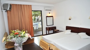 A hotel room with a bed, a dresser, a mirror, a vase with flowers, and a view of the outdoors. - Santa Marina Hotel Apartments (Kos)