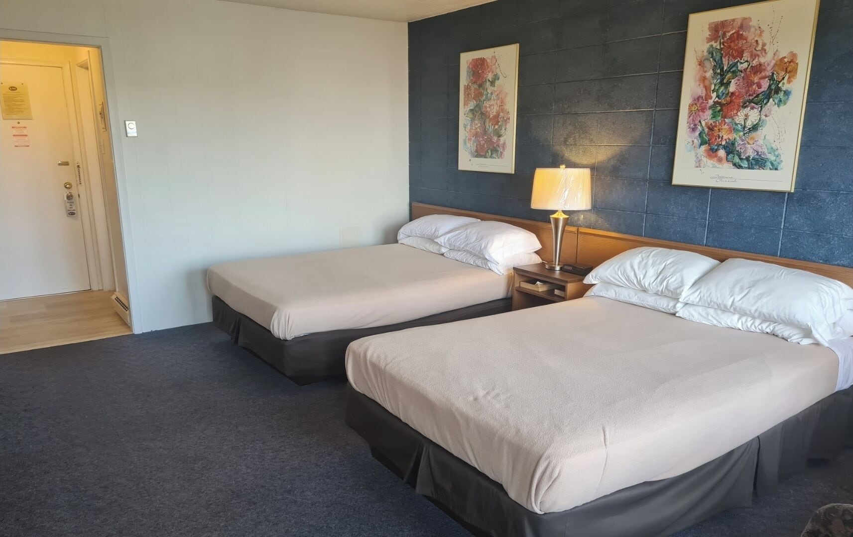 Una habitación de hotel con dos camas, una mesita de noche con lámpara y una puerta con interruptor de luz.