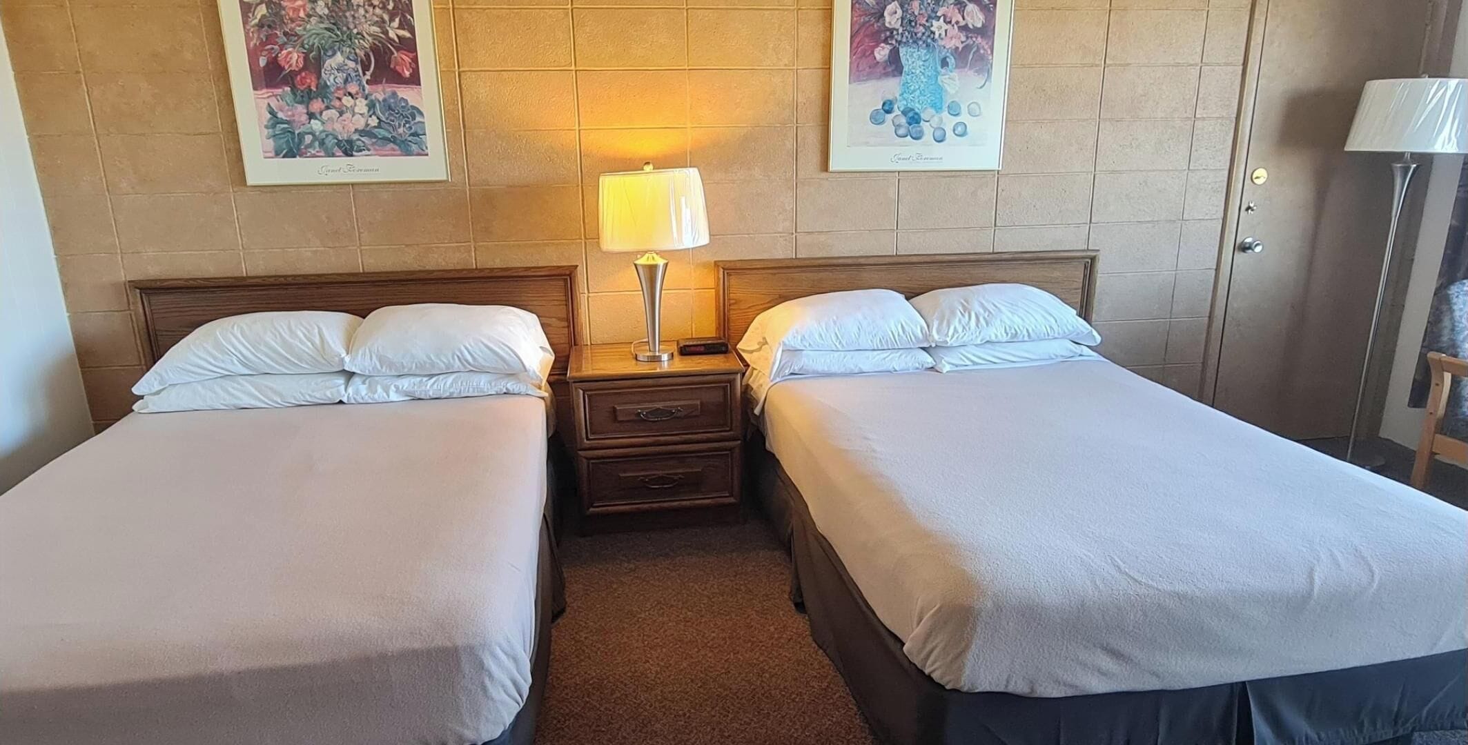 Et hotelværelse med to enkeltsenge, et natbord med lampe, og to blomsterbilleder i rammer på væggen.