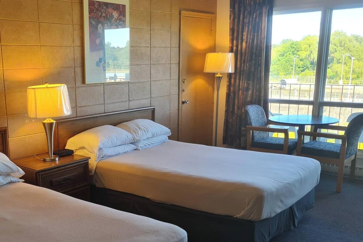 A hotel room with two beds, a nightstand, a lamp, a chair, a table, and a window with a view of greenery.