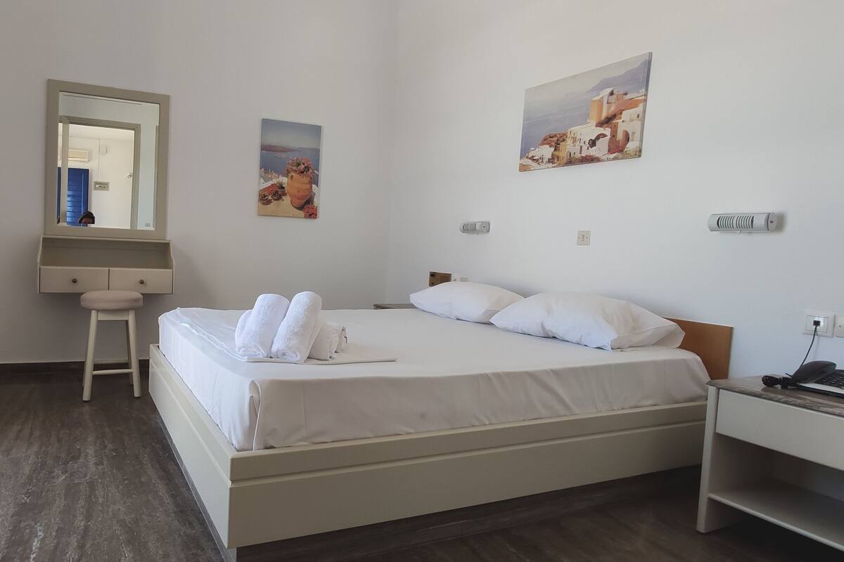 A hotel room with a bed, a nightstand, a desk, a chair, a mirror, and two paintings on the wall.