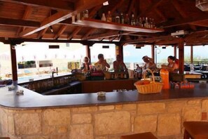 Una zona de bar con mostrador de piedra, vigas de madera y vistas a la piscina y edificios al fondo.