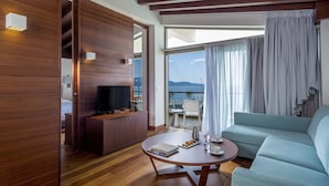 Une chambre d’hôtel moderne avec un plancher en bois, une table basse ronde sur laquelle sont posés des livres et une assiette de pâtisseries, un canapé bleu clair, une télévision et un balcon avec vue sur la mer.