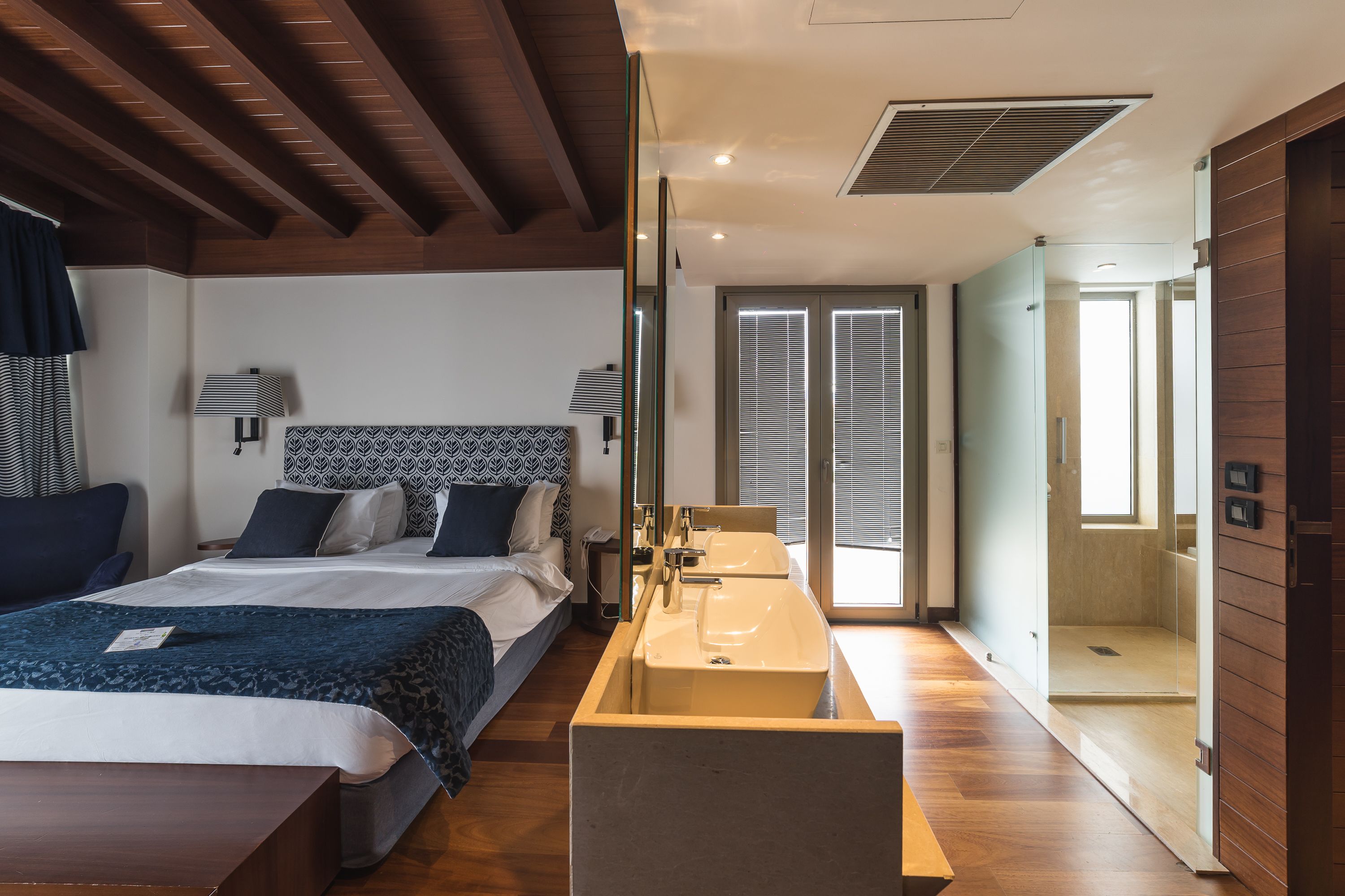 A modern hotel room with a large bed, bedside lamps, a wooden floor, a built-in sink, and a shower area.