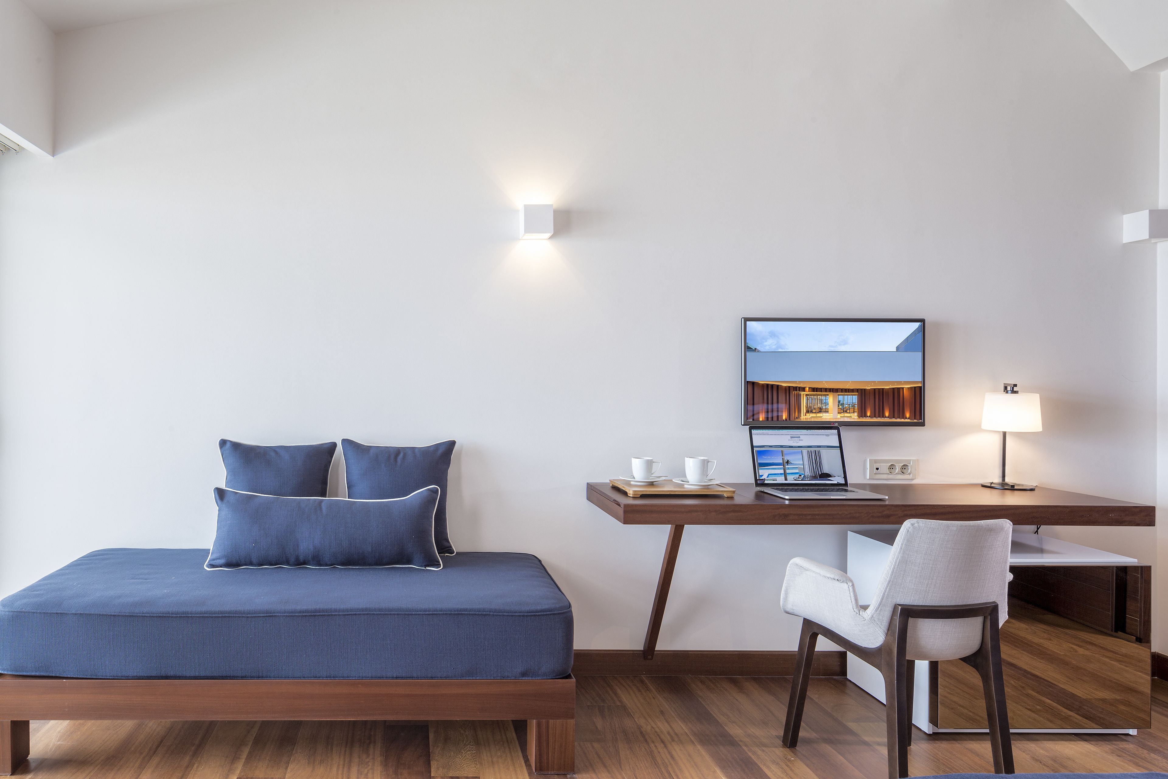 A modern bedroom with a bed, a desk with a laptop, a chair, and a TV on the wall.