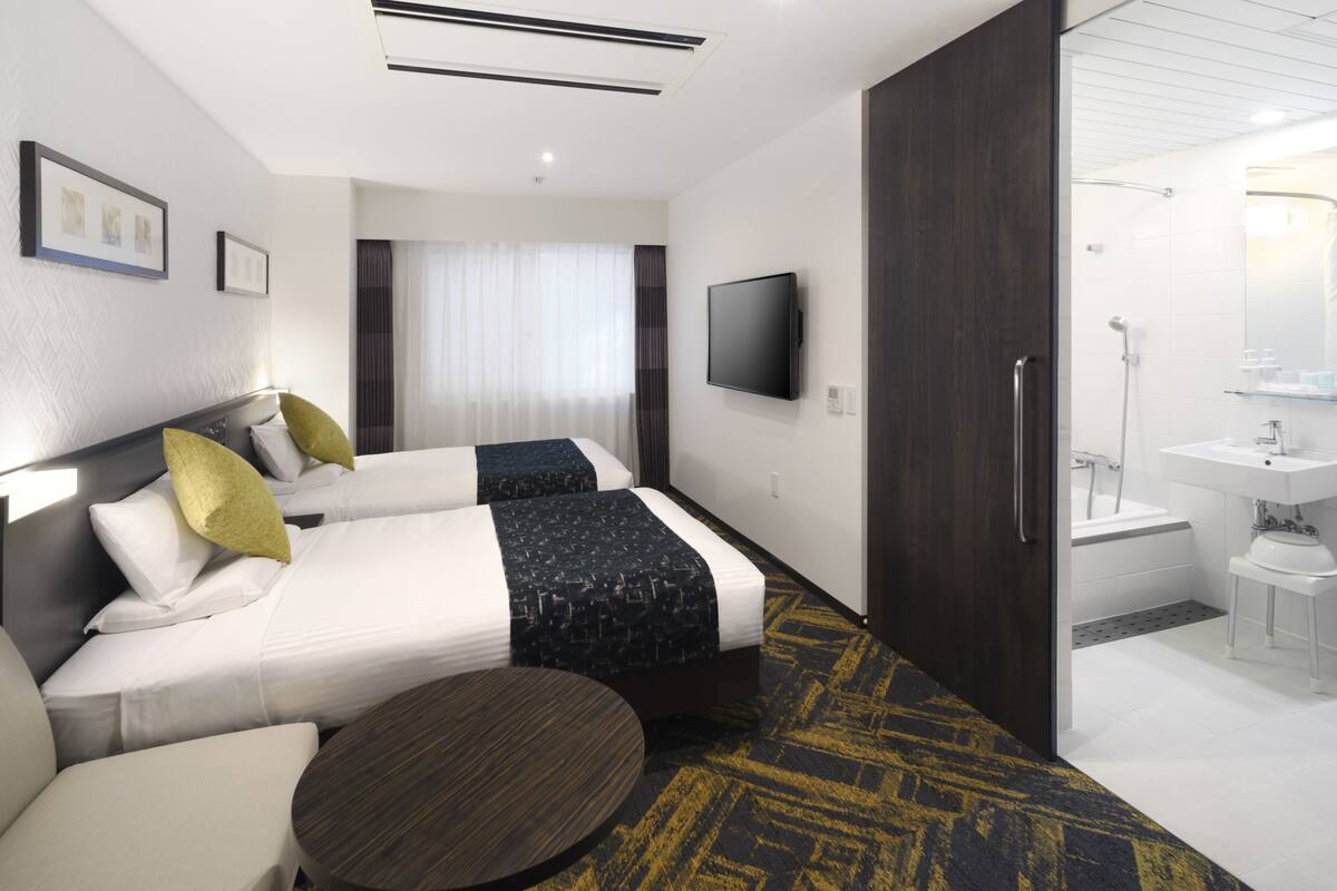 A hotel room with two beds, a round table, a TV, a bathroom with a sink and mirror, and a large window with curtains.