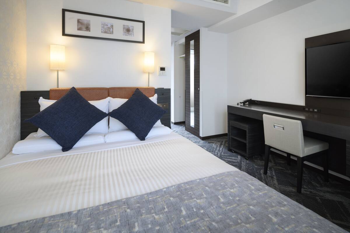 A hotel room with a bed, two pillows, a desk, a chair, a flat-screen TV, and two wall-mounted lamps.