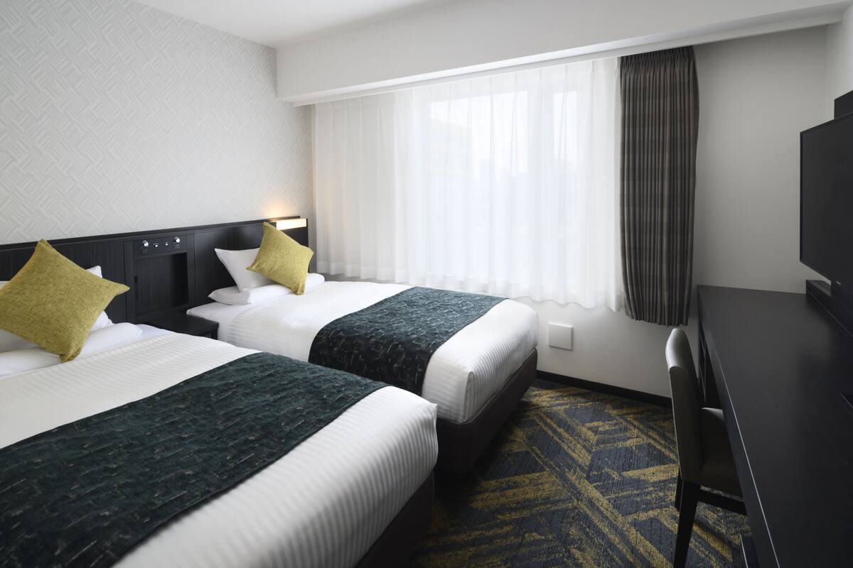 A hotel room with two beds, a desk, a television, and a window with curtains.