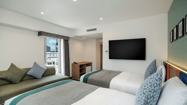 Quarto de hotel com duas camas, um sofá, uma televisão e uma janela com vista para edifícios.