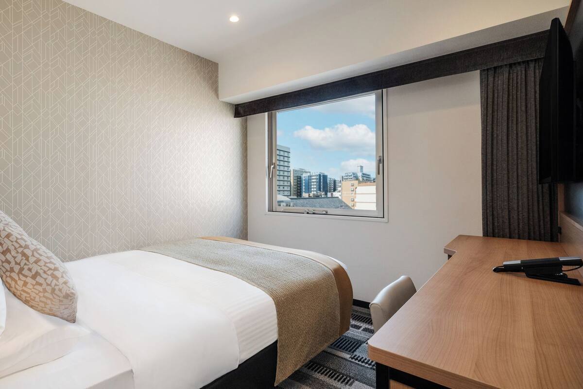 Une chambre d’hôtel équipée d’un lit, d’un bureau, d’une chaise, d’une télévision et d’une fenêtre donnant sur la ville.