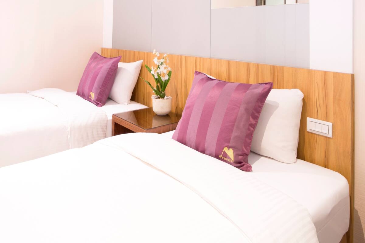 A hotel room with two beds, each with white bedding and purple pillows. There is a small table with a vase of flowers between the beds.