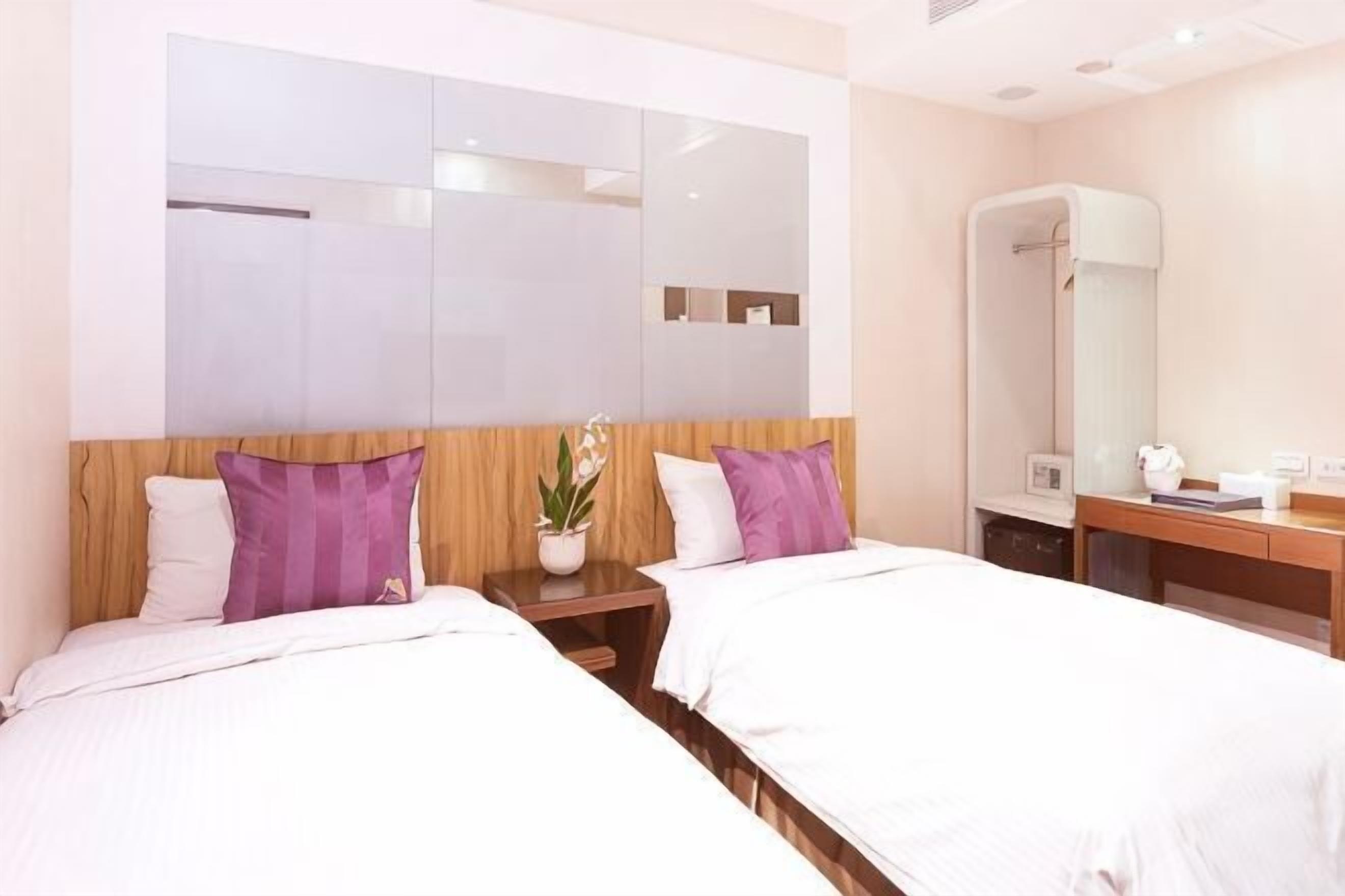 Ein Hotelzimmer mit zwei Betten, einem Holzkopfstück, einem großen Spiegel, einem Nachttisch mit Lampe sowie einem Badezimmer mit Dusche und Toilette.