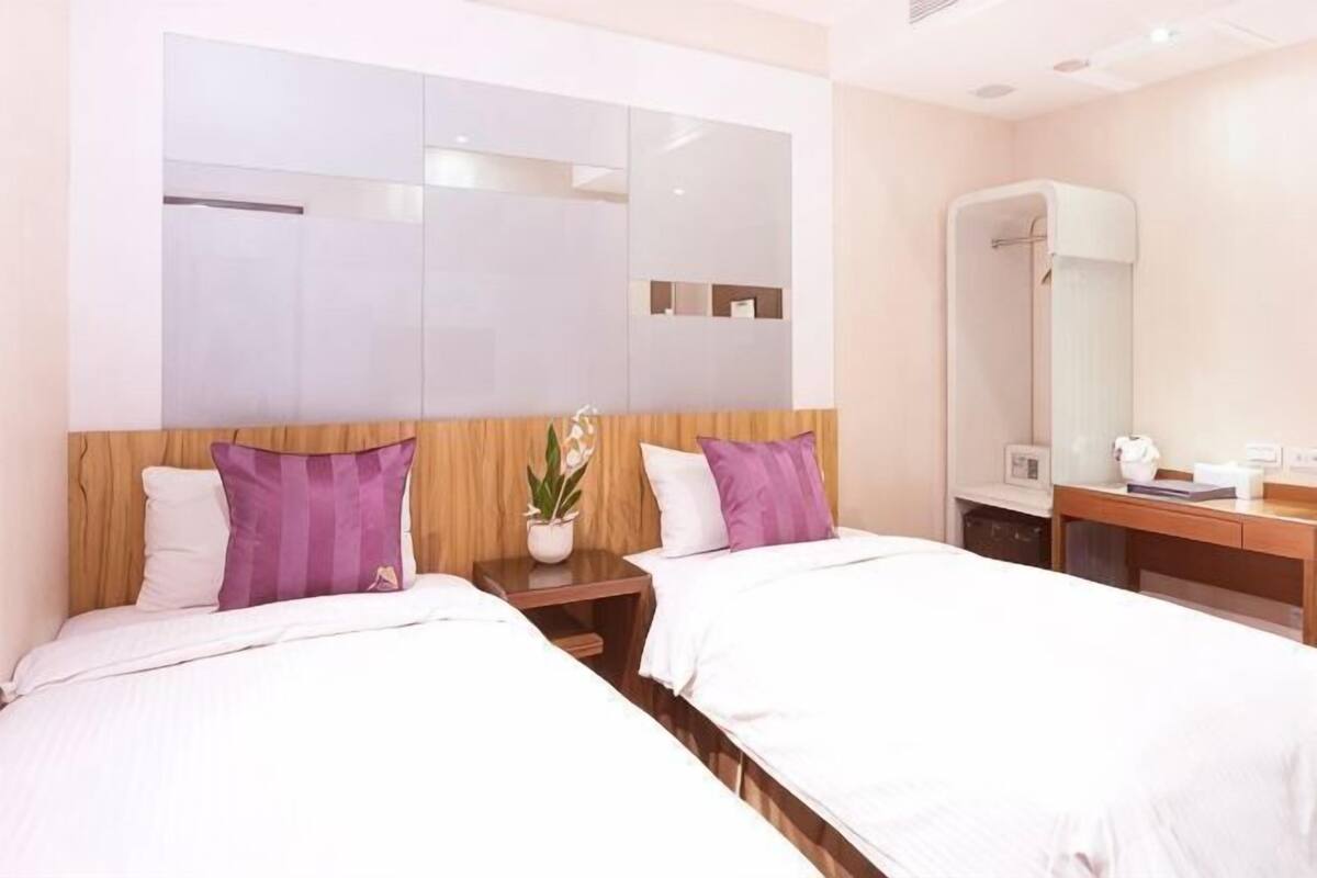 A hotel room with two beds, a wooden headboard, a large mirror, a nightstand with a lamp, and a bathroom with a shower and toilet.