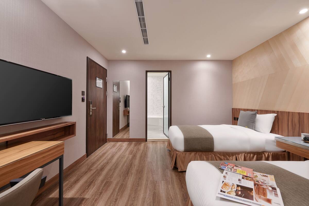 A hotel room with a large flat-screen TV, two beds, a wooden desk, and a glass shower door.