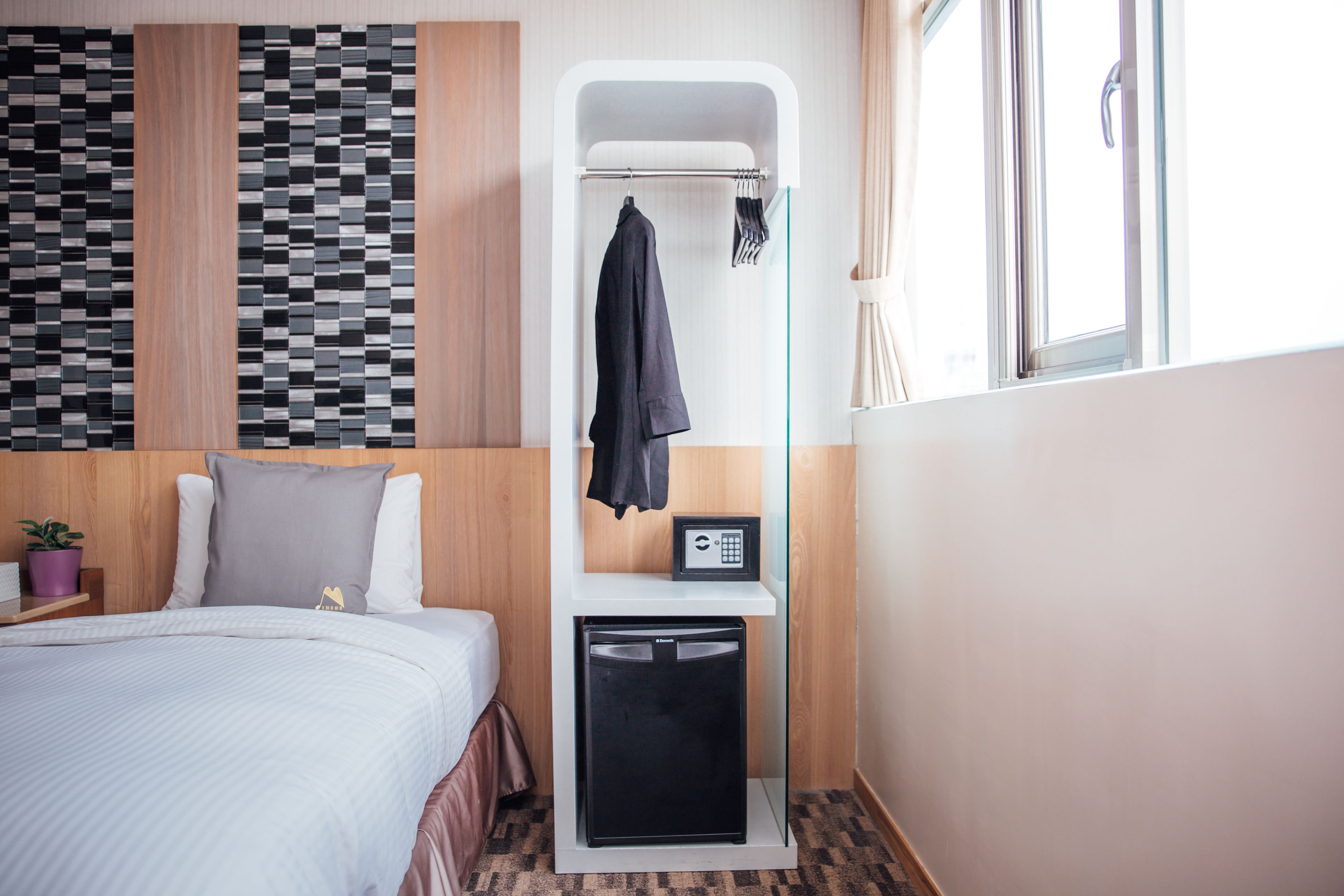 Une chambre d’hôtel comprenant un lit, un petit bureau avec un réfrigérateur, un porte-manteau fixé au mur et une fenêtre avec des rideaux.