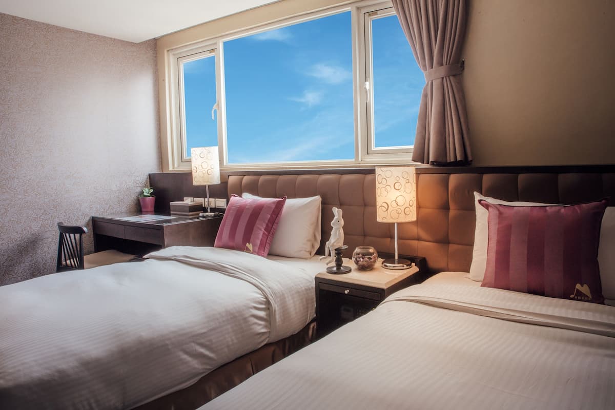 A hotel room with two beds, a nightstand, a lamp, and a window with curtains.
