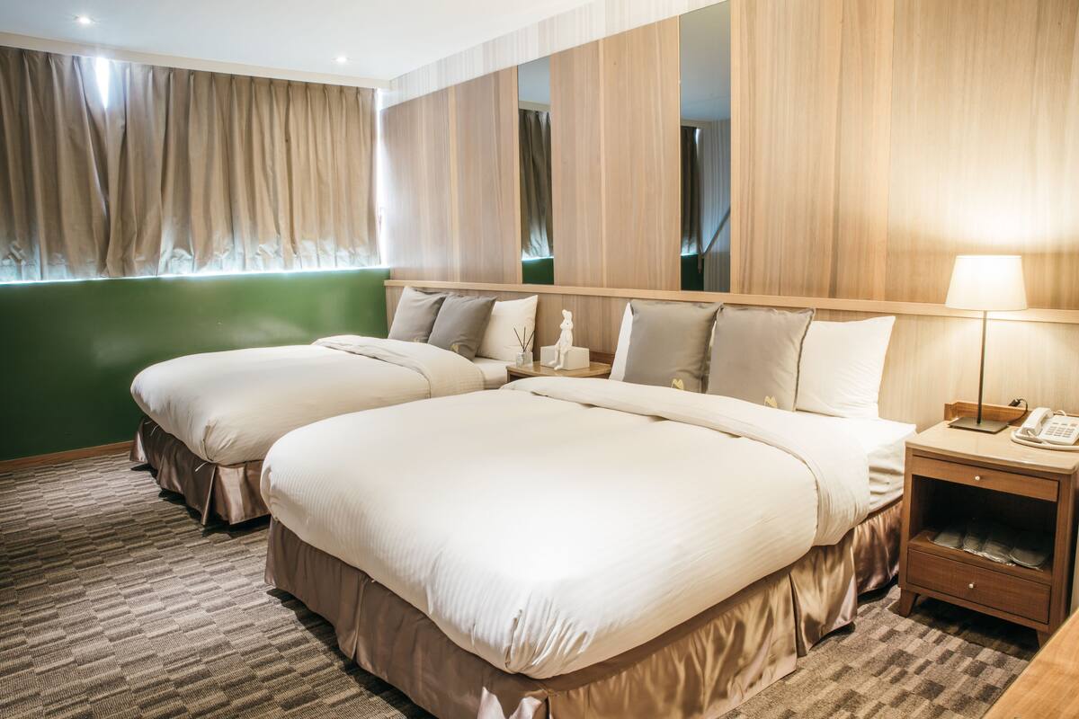 A hotel room with two beds, a green accent wall, a wooden headboard, a nightstand with a lamp, and a telephone.