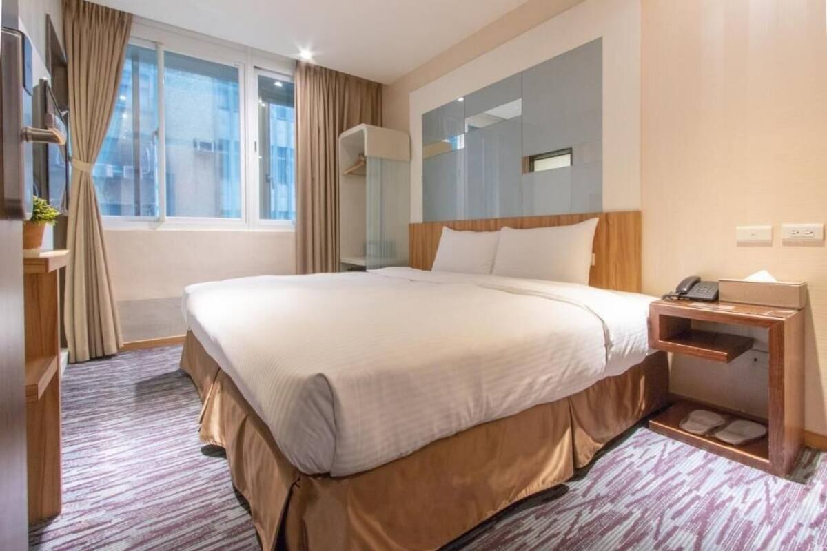 A hotel room with a large bed, a bedside table with a phone, a mirror, and a window with curtains.