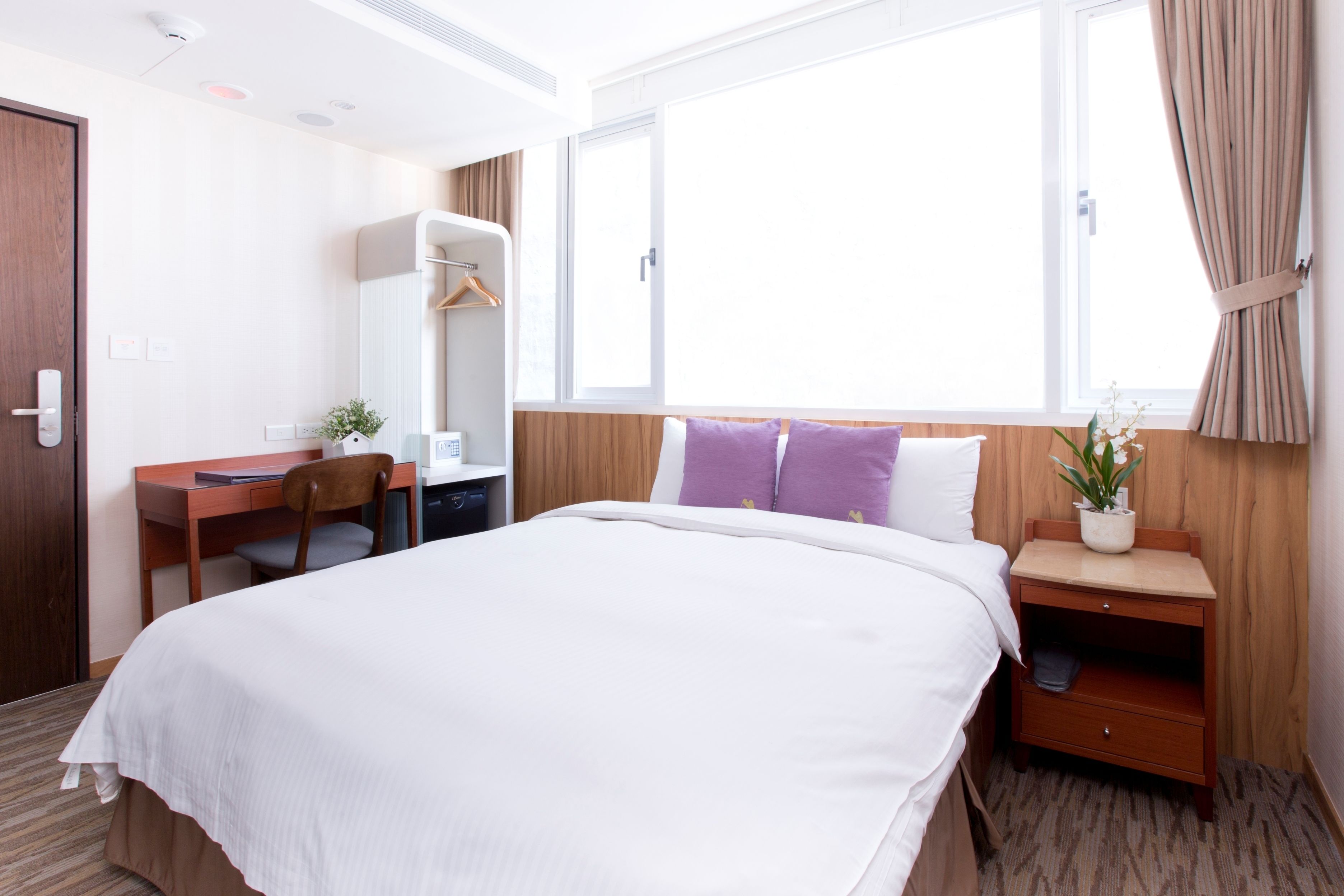 A hotel room with a bed, a desk, a chair, a wardrobe, a nightstand with a plant, and a window with curtains.