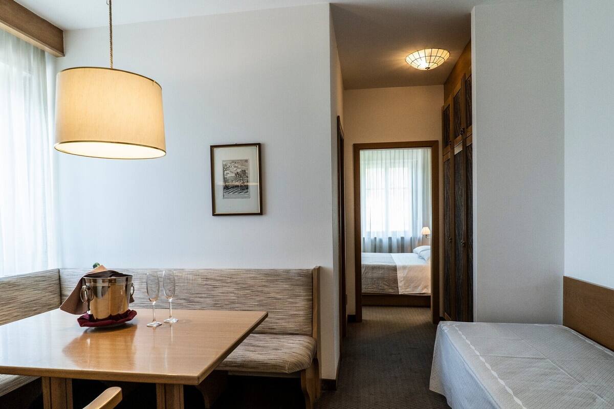 Een hotelkamer met een houten eettafel, een bankje, een bed met witte dekbedovertrek, een ingelijste foto aan de muur, en een gang naar een andere kamer.