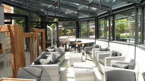 A modern outdoor lounge area with wicker furniture, glass ceiling, and large windows offering views of greenery. - Hotel Villa Regina (Levico Terme)