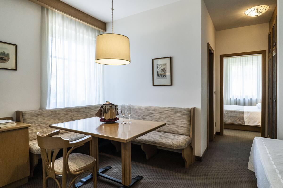 Een hotelkamer met een houten tafel, een bankje, een bed en een groot raam met gordijnen.