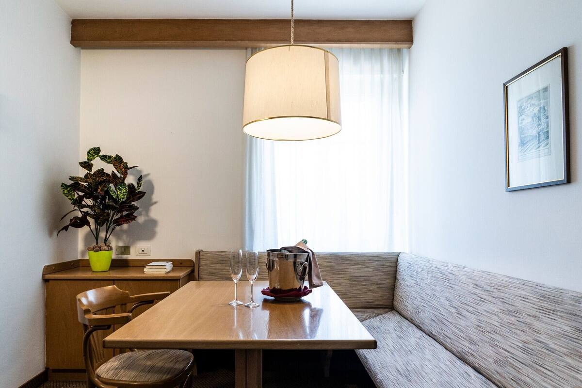 Een eetgedeelte met een houten tafel, twee stoelen, een bank, een potplant, een hanglamp en een lijst met een afbeelding aan de muur.