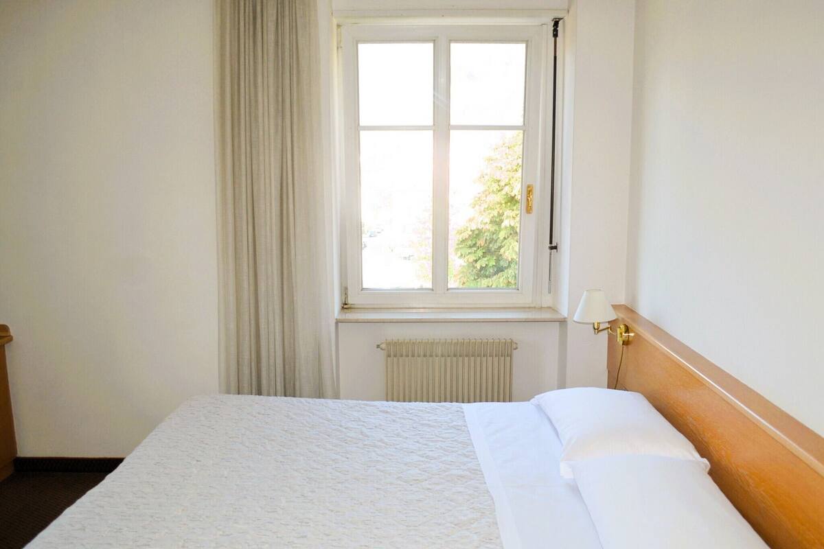 Een hotelkamer met een enkel bed, een raam met gordijnen, een radiator en een wandlamp.