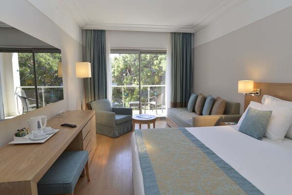 A hotel room with a bed, a desk, a chair, a small table, and a balcony with a view of trees.