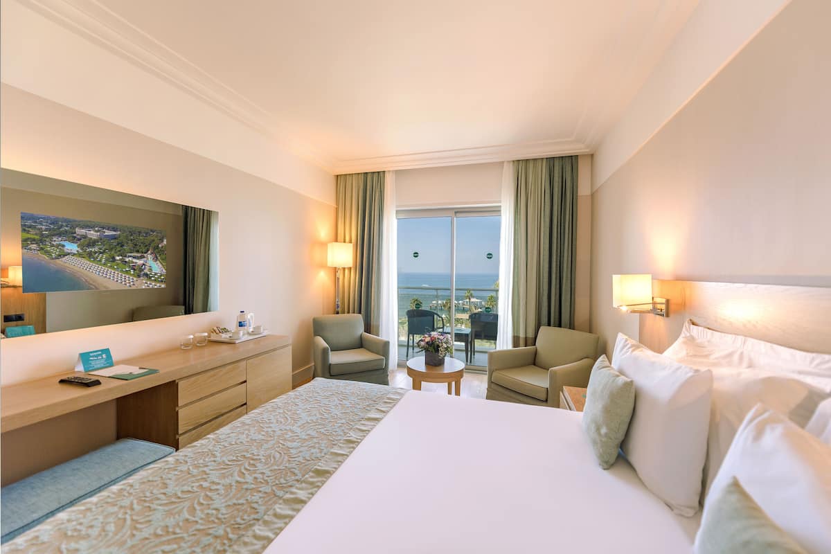 Habitación de hotel con una cama grande, un escritorio, dos sillones, una mesita con un jarrón y un balcón con vistas al mar.