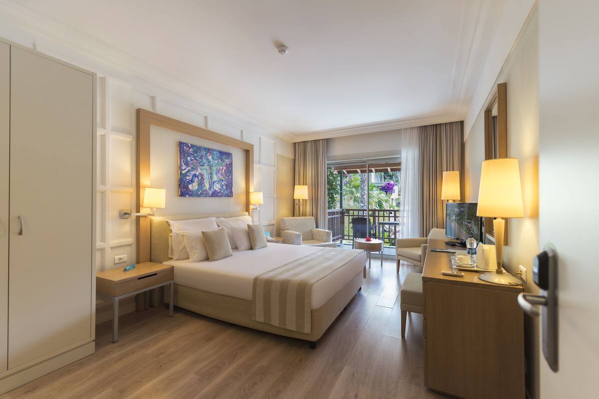 Habitación de hotel con una cama grande, mesitas de noche, un escritorio con silla y un balcón con vistas.
