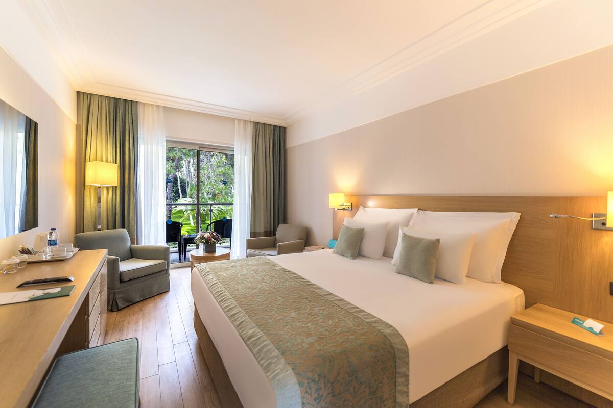 Habitación de hotel con una cama grande, un escritorio con silla, una mesita con silla y vistas al exterior a través de una puerta de vidrio.