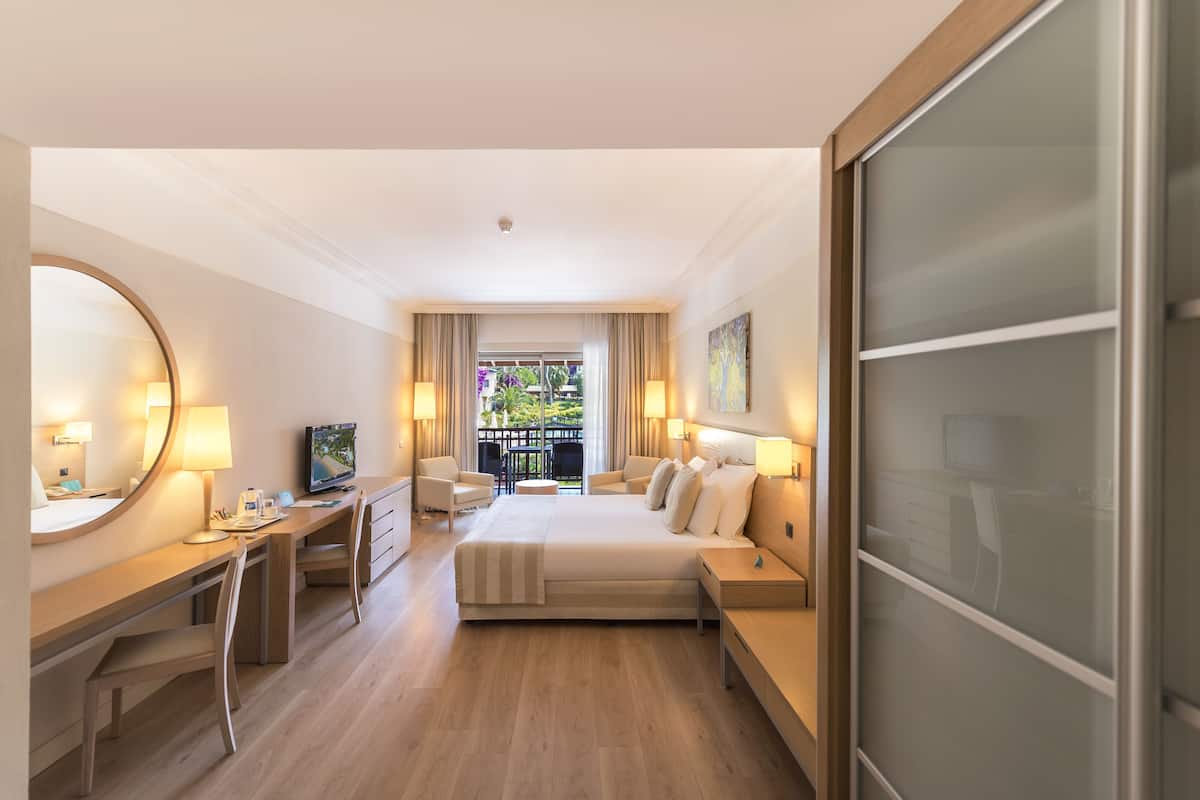 Una habitación de hotel moderna con una cama grande, un escritorio, una silla, un televisor y un balcón con vistas a zonas verdes.