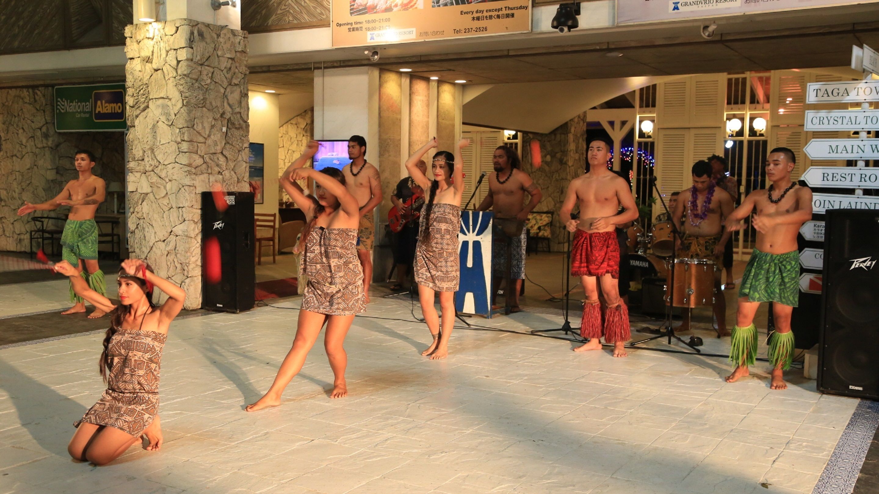 A group performing on a stage with a stone pillar and a sign indicating the resort's operating hours.