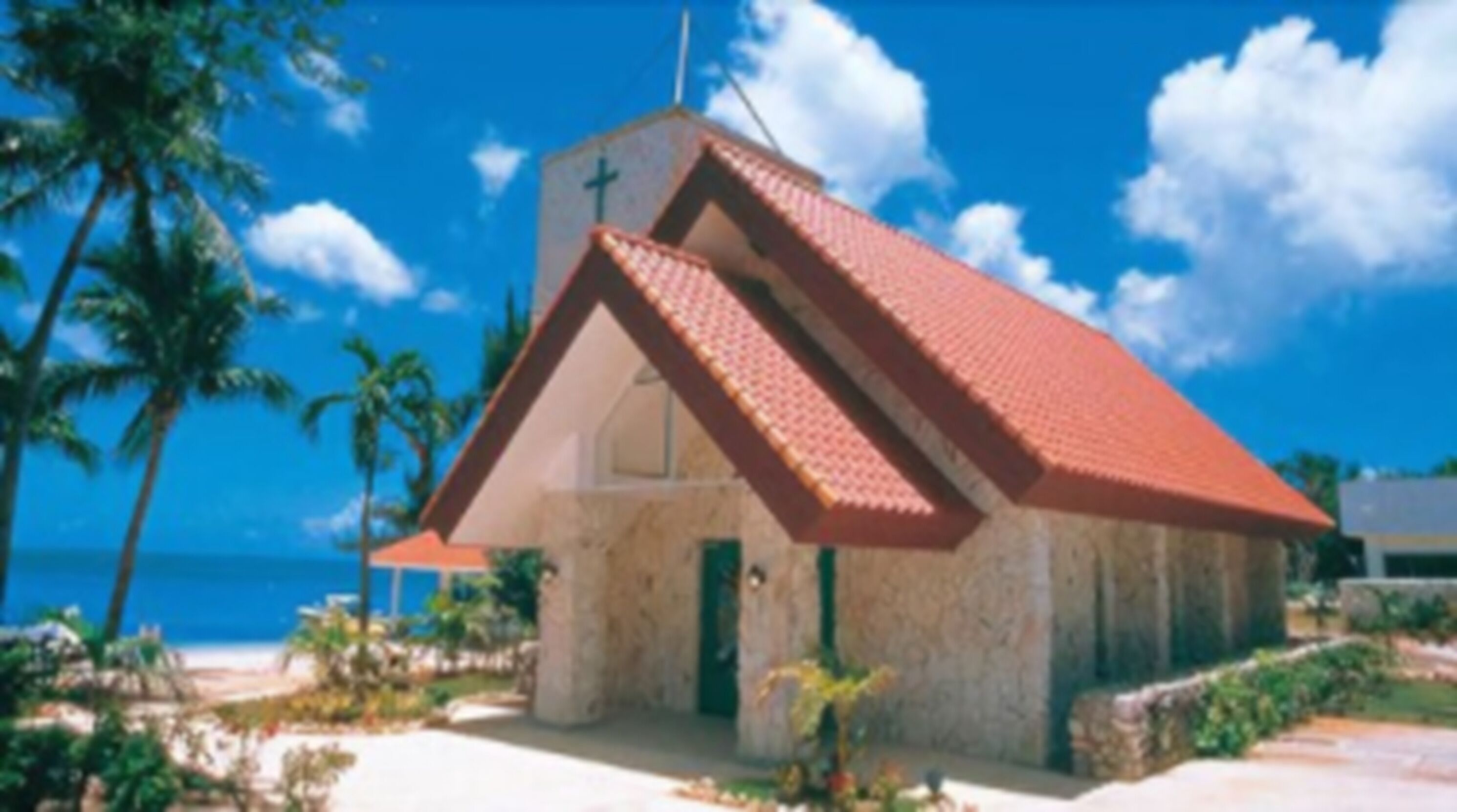 A small church with a red roof and a cross on top, surrounded by palm trees and a clear blue sky.