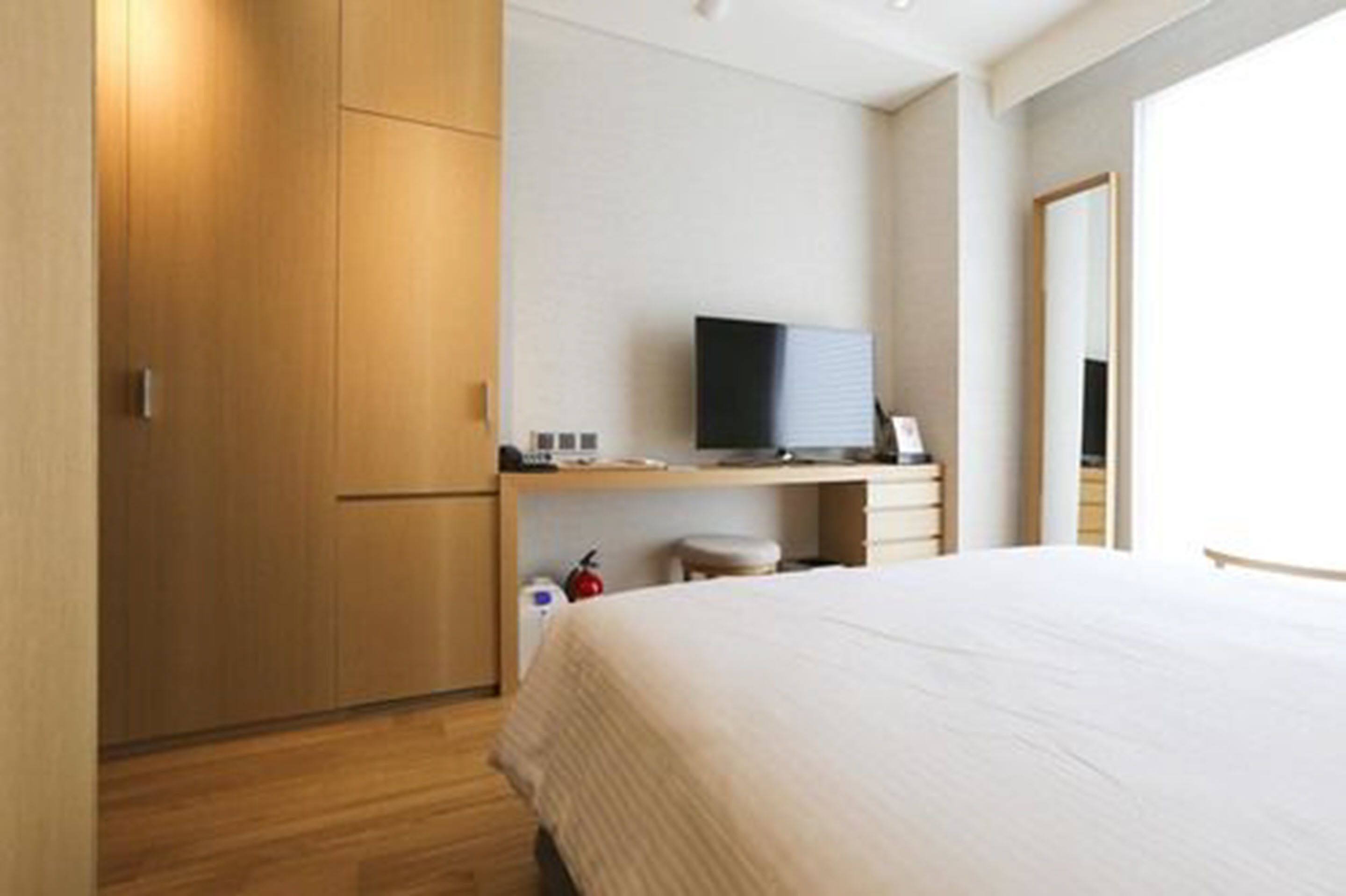 A hotel room with a bed, a desk with a TV, a wardrobe, and a small stool.
