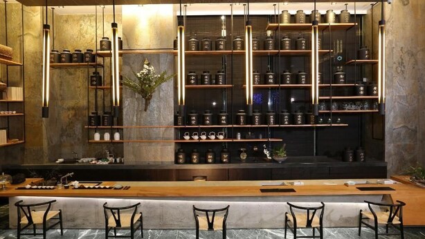 Uma área de bar com prateleiras de garrafas e copos, um balcão de madeira e banquetas.