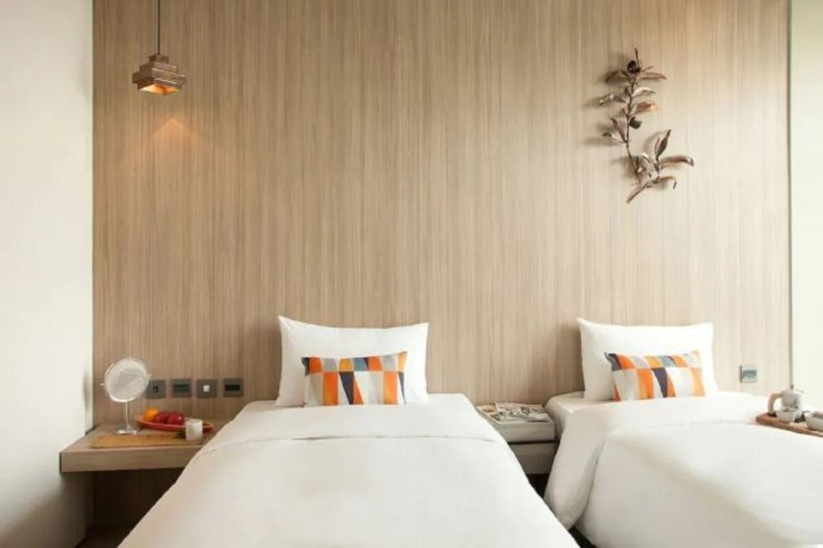 A hotel room with two beds, a wooden headboard, a small bedside table with a mirror, and a decorative plant.